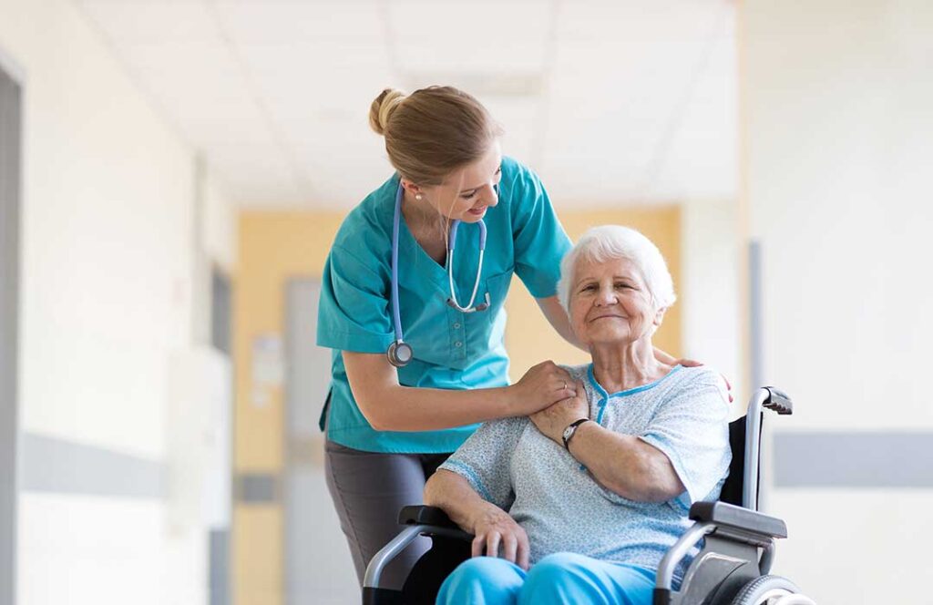 Two older adults sitting at a dining table with a caregiver serving breakfast in a sunlit community room.